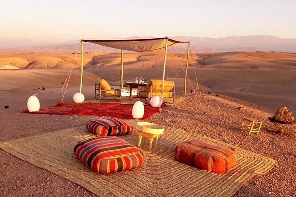 Luxury Agafay desert glamping with beige canopy, red Moroccan cushions, ambient lighting, and rolling desert dunes at golden hour